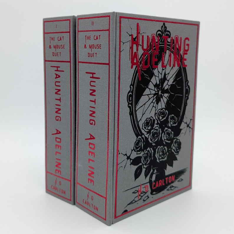 May include: Two hardcover books titled "Hunting Adeline" and "The Cat & Mouse Duet". The books have gray covers with red text and accents. The cover art features a black and white illustration of a broken mirror and roses.