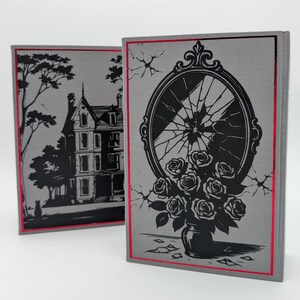 May include: Two hardcover books with gray covers and red edges. One book features a black illustration of a house and tree. The other book has a black illustration of a broken mirror and a vase of roses.
