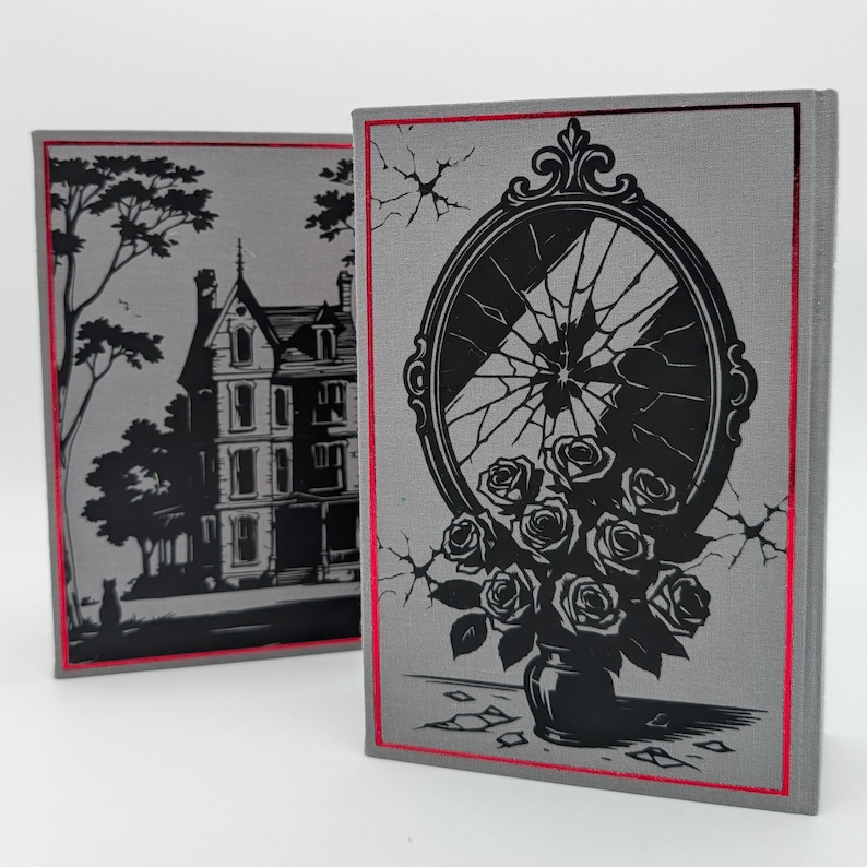 May include: Two hardcover books with gray covers and red edges. One book features a black illustration of a house and tree. The other book has a black illustration of a broken mirror and a vase of roses.