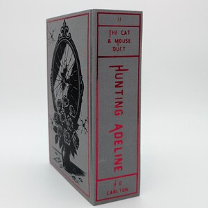 May include: A hardcover book titled "Hunting Adeline" with a gray cover and red accents. The cover features a black illustration of a broken mirror and roses. The spine displays the title and author's name in red.