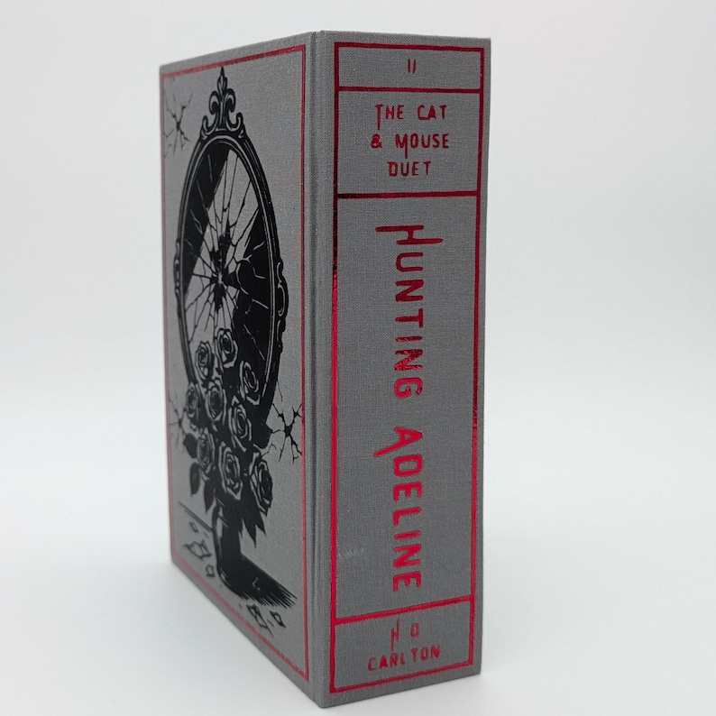 May include: A hardcover book titled "Hunting Adeline" with a gray cover and red accents. The cover features a black illustration of a broken mirror and roses. The spine displays the title and author's name in red.