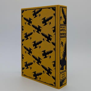 May include: A hardcover book with a mustard yellow fabric cover featuring a repeating black pattern of spaceships and astronauts. The spine displays the title Project Hail Mary and the author name Andy Weir in black text within rectangular panels.