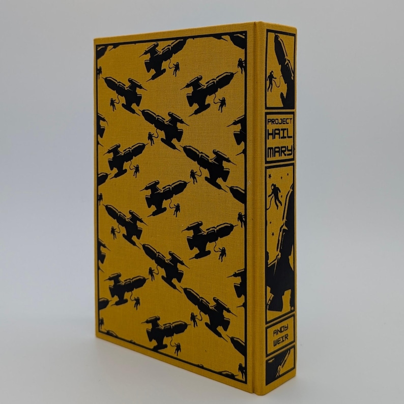 May include: A hardcover book with a mustard yellow fabric cover featuring a repeating black pattern of spaceships and astronauts. The spine displays the title Project Hail Mary and the author name Andy Weir in black text within rectangular panels.