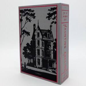 May include: A hardcover book with a gray cover featuring a black illustration of a house and trees. The spine is red with the title "Haunting Adeline" and the author's name. The book is a literary work.
