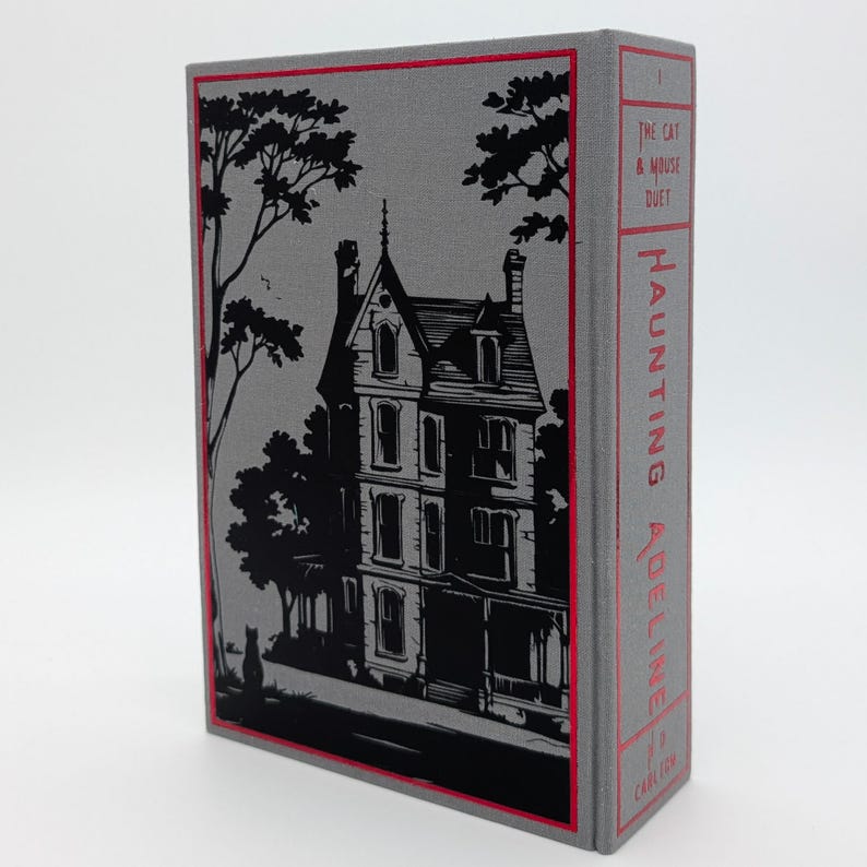 May include: A hardcover book with a gray cover featuring a black illustration of a house and trees. The spine is red with the title "Haunting Adeline" and the author's name. The book is a literary work.