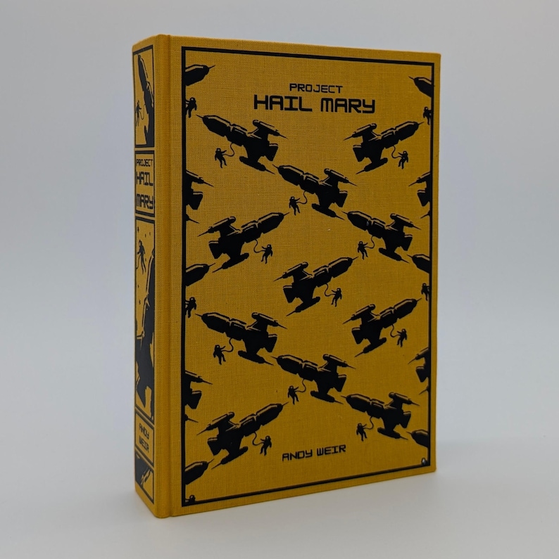 May include: A hardcover book titled Project Hail Mary by Andy Weir with a mustard yellow fabric cover. The front features a repeating black pattern of spaceships and astronauts. The spine includes the title and author name in black text.