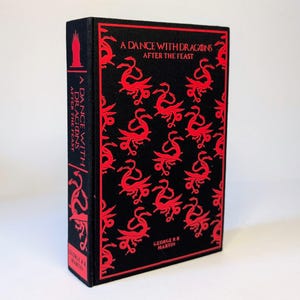 May include: A hardcover book titled "A Dance with Dragons: After the Feast" by George R.R. Martin. The book has a black cover with a repeating red dragon pattern and red text. The spine also features the title and author.