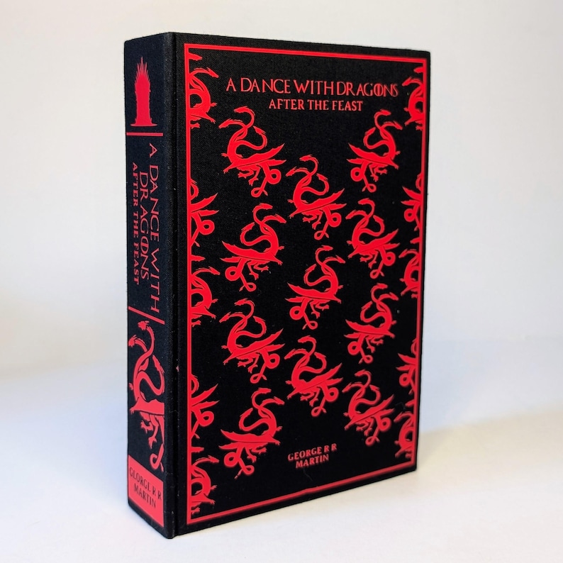 May include: A hardcover book titled "A Dance with Dragons: After the Feast" by George R.R. Martin. The book has a black cover with a repeating red dragon pattern and red text. The spine also features the title and author.