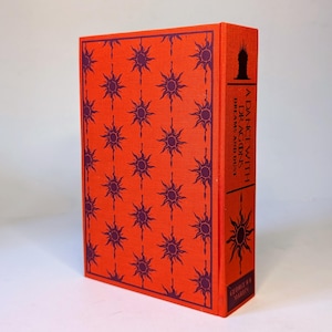 May include: A hardcover book titled "A Dance with Dragons Dreams and Dust" by George R.R. Martin. The book has a vibrant orange cover with a repeating pattern of purple sunburst designs. The spine features the title and author's name.