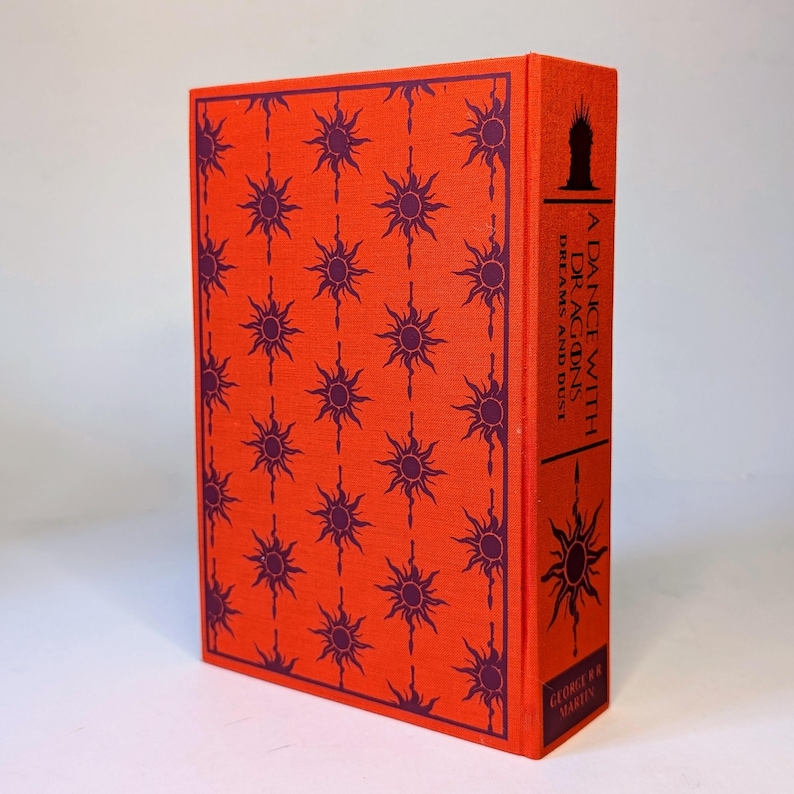 May include: A hardcover book titled "A Dance with Dragons Dreams and Dust" by George R.R. Martin. The book has a vibrant orange cover with a repeating pattern of purple sunburst designs. The spine features the title and author's name.