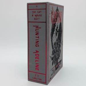 May include: A hardcover book titled "Hunting Adeline" with a gray cover and red accents. The spine features the title and author's initials. The front cover displays a black illustration of a mirror with roses. The book is part of "The Cat & Mouse Duet" series.