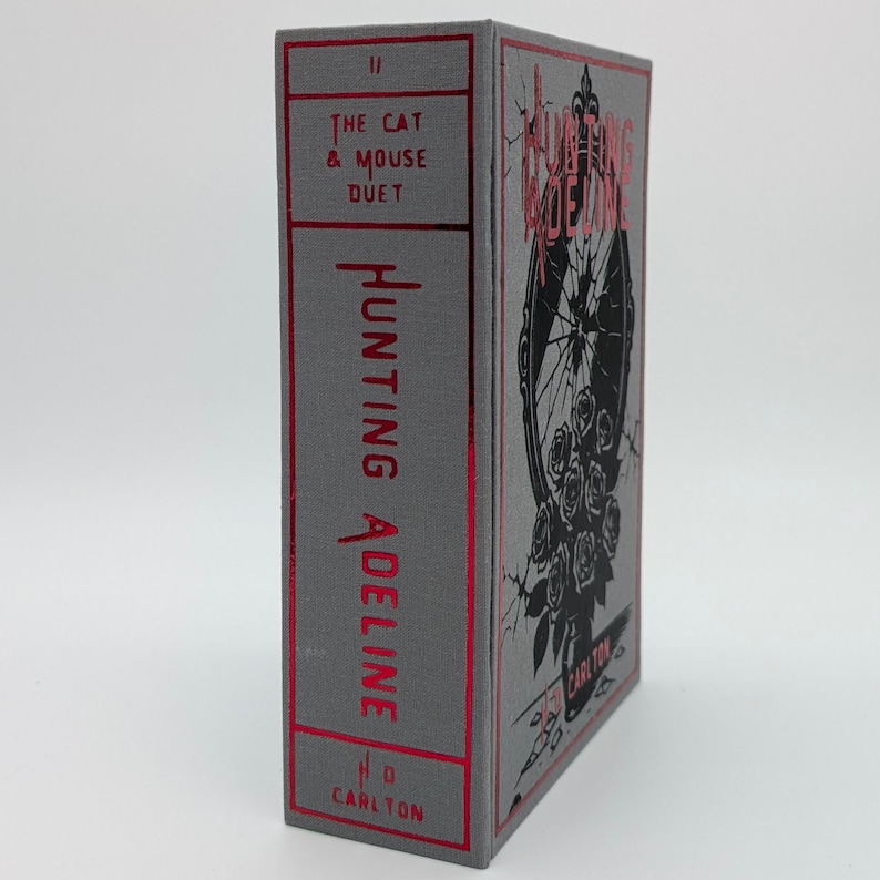 May include: A hardcover book titled "Hunting Adeline" with a gray cover and red accents. The spine features the title and author's initials. The front cover displays a black illustration of a mirror with roses. The book is part of "The Cat & Mouse Duet" series.