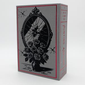 May include: A hardcover book with a gray cover featuring a black illustration of a broken mirror and a vase of roses. The title "Hunting Adeline" is printed vertically on the spine in red, with the words "The Cat & Mouse Duet" above it.