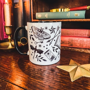 May include: White ceramic mug with a black rim and handle. The mug features a black and white design of a moth, a key, a bat, an hourglass, arrows, a star, and leaves.