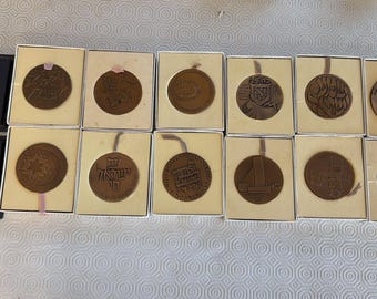 Puede incluir: Una colección de medallas de color bronce, cada una en cajas individuales. Las medallas presentan varios diseños, incluyendo texto, símbolos y patrones geométricos. Algunas están en cajas negras, otras en cajas color crema.