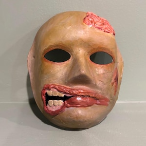 May include: A grotesque mask with a pale complexion, open eye sockets, and a gaping mouth revealing teeth and a tongue. The mask has a wound on the forehead and cheek, with red and pink accents. The mask is likely for Halloween or costume use.