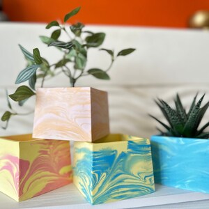May include: Three square-shaped planters with a marbled design. The planters are in shades of yellow, pink, and blue. The planter on the top is peach with white swirls, the planter on the left is yellow with pink swirls, and the planter on the right is blue with yellow swirls.
