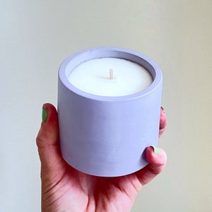 May include: A lilac-coloured candle in a cylindrical concrete container. The candle has a white wax surface and a single, central wick. The container is held in a hand, showing the candle's size and design.