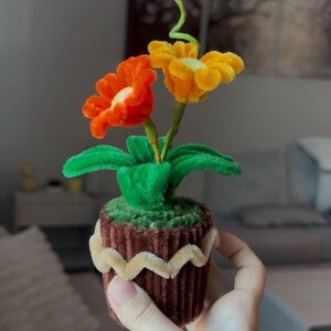 Pode incluir: Um objeto decorativo artesanal com duas flores de limpador de cachimbo, uma laranja e uma amarela, com folhas e caule verdes. As flores estão fixadas numa base cilíndrica castanha canelada, decorada com um rebordo em ziguezague bege.