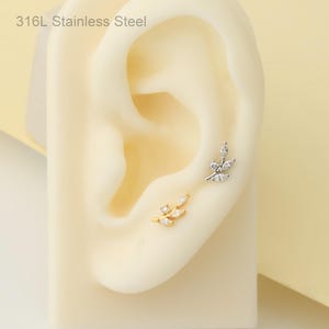May include: Two leaf-shaped earrings, one gold-coloured and one silver-coloured, each set with clear gemstones. The earrings are shown on a beige ear model. The text "316L Stainless Steel" is visible in the upper left corner.