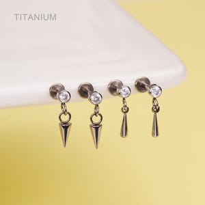 May include: Four titanium earrings with a small, clear gem at the top and a dangling silver-colored charm. Two earrings have a cone-shaped charm, and two have a teardrop-shaped charm. The word "Titanium" is printed in the upper left corner.