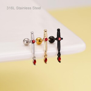May include: Three stainless steel sword-shaped earrings in silver, gold, and black. Each earring features a red gem at the hilt and a dangling red gem at the tip. The text "316L Stainless Steel" is visible.