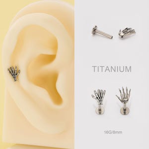 May include: Silver-colored titanium ear piercings featuring a skeleton hand design. The image includes a close-up of an ear with a piercing, along with additional piercing options. The text "TITANIUM" and "16G/8mm" are also visible.