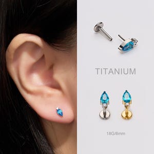 Può includere: Gioielli per piercing all'orecchio con una gemma blu chiaro a forma di goccia e una piccola pietra d'accento trasparente. La parola "TITANIUM" è stampata nell'immagine. I gioielli sono 18G/6mm.