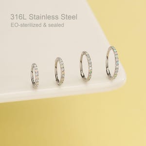 May include: Four silver hoop earrings with opal and clear crystal accents are displayed on a white surface against a yellow background. The text "316L Stainless Steel EO-sterilized & sealed" is visible.