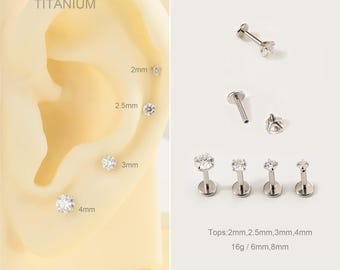 May include: A collection of titanium ear studs with clear, round gemstones. Sizes range from 2mm to 4mm. The image includes a display of the studs on an ear model and individual studs. Text on the image reads "TITANIUM".