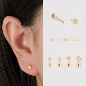 Puede incluir: Pendientes de botón y labrets texturizados de color dorado. La imagen muestra un primer plano de una oreja con un pendiente de botón y varios pendientes con el texto "14K SOLID GOLD".