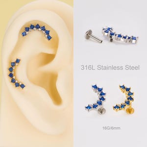 Op de afbeelding: Oorpiercings met blauwe vierkante edelstenen en kleine heldere edelstenen. De piercings hebben een gebogen vorm. De tekst "316L Stainless Steel" is zichtbaar. De afbeelding toont ook een piercing met een maat van 16G/6mm.