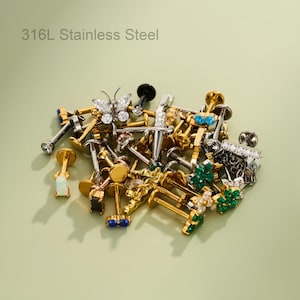 Puede incluir: Una colección de joyería corporal de acero inoxidable 316L, que incluye pendientes labret con varios adornos como formas de mariposa, daga y flor, con gemas transparentes, verdes, azules y negras en tonos dorados y plateados.