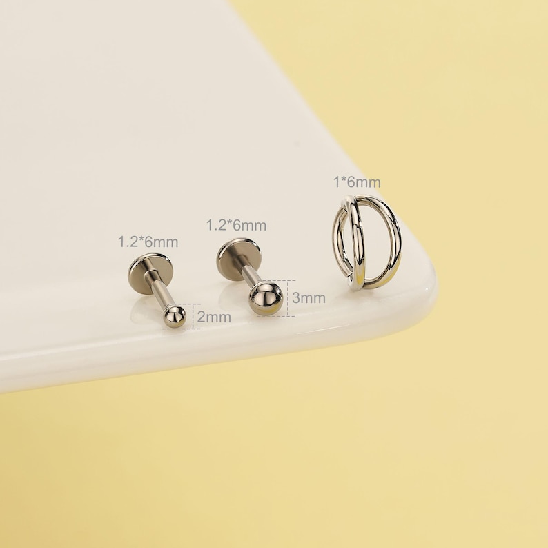 Pode incluir: Uma cole&ccedil;&atilde;o de joias corporais prateadas, incluindo dois labrets de costas planas com extremidades esf&eacute;ricas e um brinco de argola dupla. Os labrets medem 1,2 mm x 6 mm, com extremidades esf&eacute;ricas de 2 mm e 3 mm de di&acirc;metro. O brinco de argola dupla mede 1 mm x 6 mm.