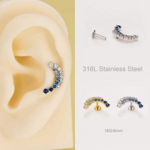 May include: A curved ear piercing stud with a gradient of white and blue gemstones. The stud is made of 316L stainless steel. The image also shows a separate stud and two additional studs with a 6mm length.