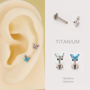 May include: Butterfly-shaped titanium earrings with colorful gemstones. The image shows the earrings in a variety of styles, including a flat-back stud and a labret stud. The text "TITANIUM" and size information are also visible.
