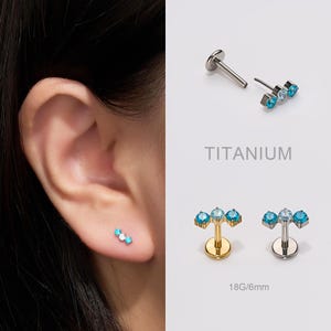 Può includere: Gioielli per piercing all'orecchio in titanio con tre pietre preziose blu. L'immagine mostra i gioielli in uso su un orecchio, e anche pezzi separati. Il testo "TITANIUM" è visibile. I gioielli sono 18G/6mm.