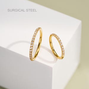 Puede incluir: Dos anillos de clic de acero quirúrgico de color dorado con una fila de piedras de circonita cúbica transparentes alrededor del borde.