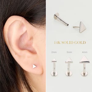 May include: A close-up of an ear with a small, triangular, silver stud earring. The image also shows three other stud earrings with triangle tops in 2mm, 3mm, and 4mm sizes. The text "14K SOLID GOLD" is visible.