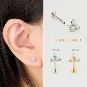 Op de afbeelding: Een oor met een kleine, witte opaal oorbel. De afbeelding toont ook een 14K massief gouden oorbel met drie opalen en een goudkleurige oorbel met drie opalen. De tekst "14K SOLID GOLD" is zichtbaar.