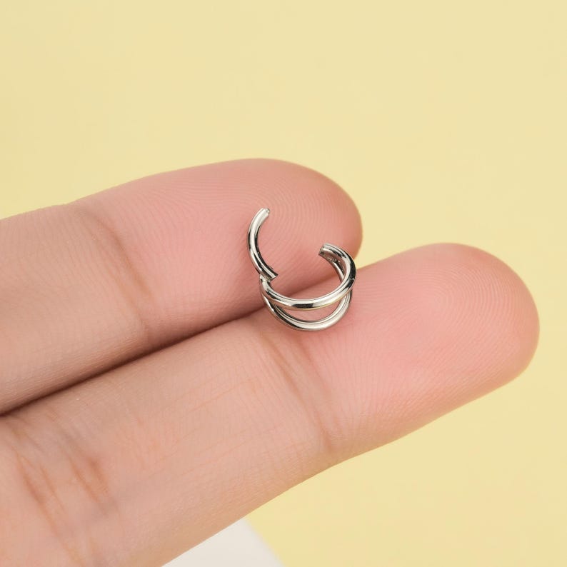 Pode incluir: Um pequeno brinco de argola prateado com um design de camada dupla. O brinco est&aacute; aberto, revelando o mecanismo de fecho. A joia &eacute; segurada contra um fundo amarelo claro.