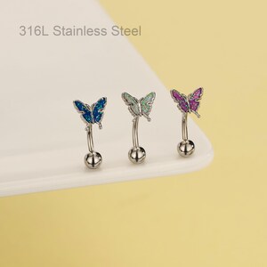 May include: Three stainless steel belly button rings with butterfly designs. Each ring features a curved barbell with a small ball at the bottom and a butterfly at the top. The butterflies are blue, green, and purple.