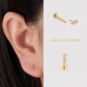 May include: A close-up of an ear with a small gold earring. The earring has a curved design. The image also shows a gold stud and a flat-backed earring. The text "14K SOLID GOLD" is displayed.