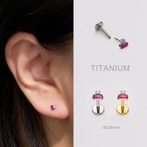May include: Titanium ear piercing studs with a rectangular pink gemstone and a small clear gem. The image shows the studs in use on an ear, and also separately. The text "TITANIUM" and "18G/6mm" are also visible.