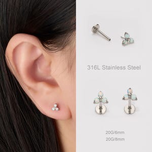 Op de afbeelding: Een close-up van een zilveren oorbel met drie opaalachtige stenen. De tekst "316L Stainless Steel" is zichtbaar. De afbeelding bevat ook de tekst "20G/6mm" en "20G/8mm".