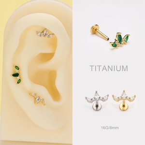 May include: An assortment of ear piercings, including a gold stud with green gems, a silver stud with clear gems, and a gold stud with clear gems. The word "TITANIUM" is printed in gray.