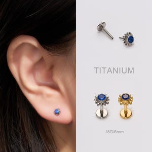 Puede incluir: Primer plano de una oreja con un pequeño pendiente de botón de piedra preciosa azul. La imagen también muestra dos pendientes de botón adicionales con un centro de piedra preciosa azul y un engaste plateado u dorado. El texto "TITANIUM" y "18G/6mm" son visibles.