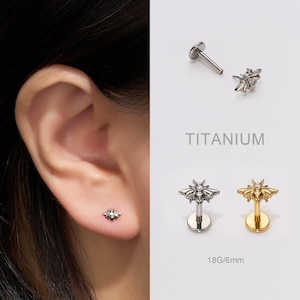 Puede incluir: Pendiente de botón con forma de abeja en tonos plateados y dorados. La imagen muestra el pendiente en una oreja y por separado. También se ve el texto "TITANIUM" y "18G/6mm".