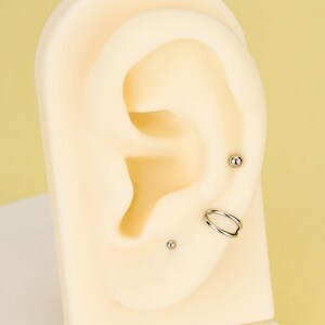 Pode incluir: Uma exibi&ccedil;&atilde;o de piercings na orelha em uma orelha de manequim. Os piercings incluem dois pequenos brincos de prata e um brinco de argola dupla. A orelha do manequim &eacute; de cor bege clara, em um fundo amarelo.