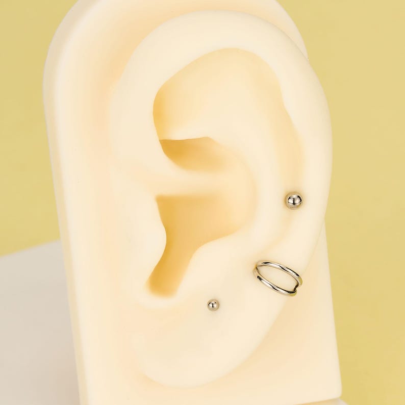 Pode incluir: Uma exibi&ccedil;&atilde;o de piercings na orelha em uma orelha de manequim. Os piercings incluem dois pequenos brincos de prata e um brinco de argola dupla. A orelha do manequim &eacute; de cor bege clara, em um fundo amarelo.